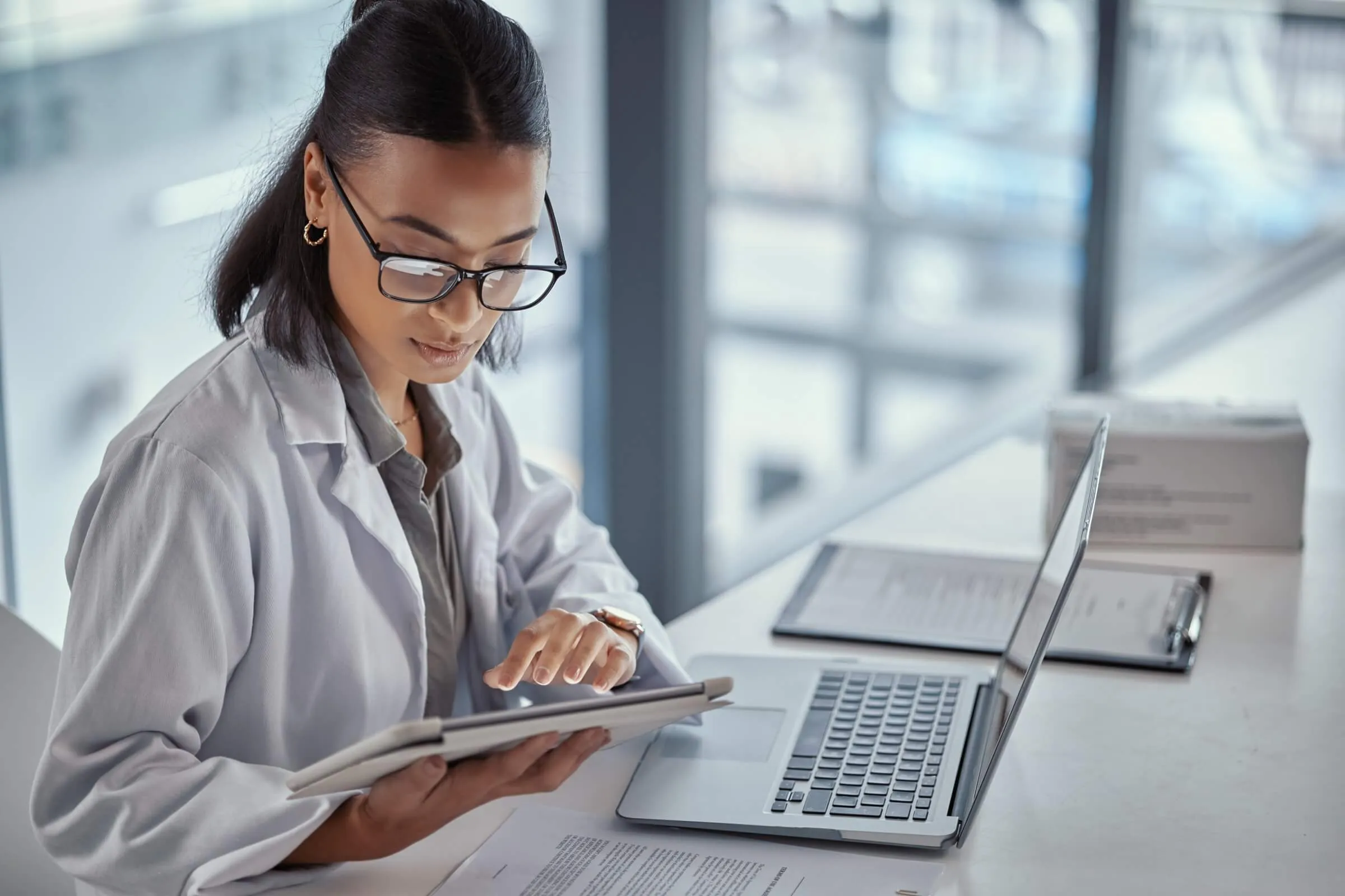 Doctor in glasses taking notes while reviewing information on laptop in bright office space.