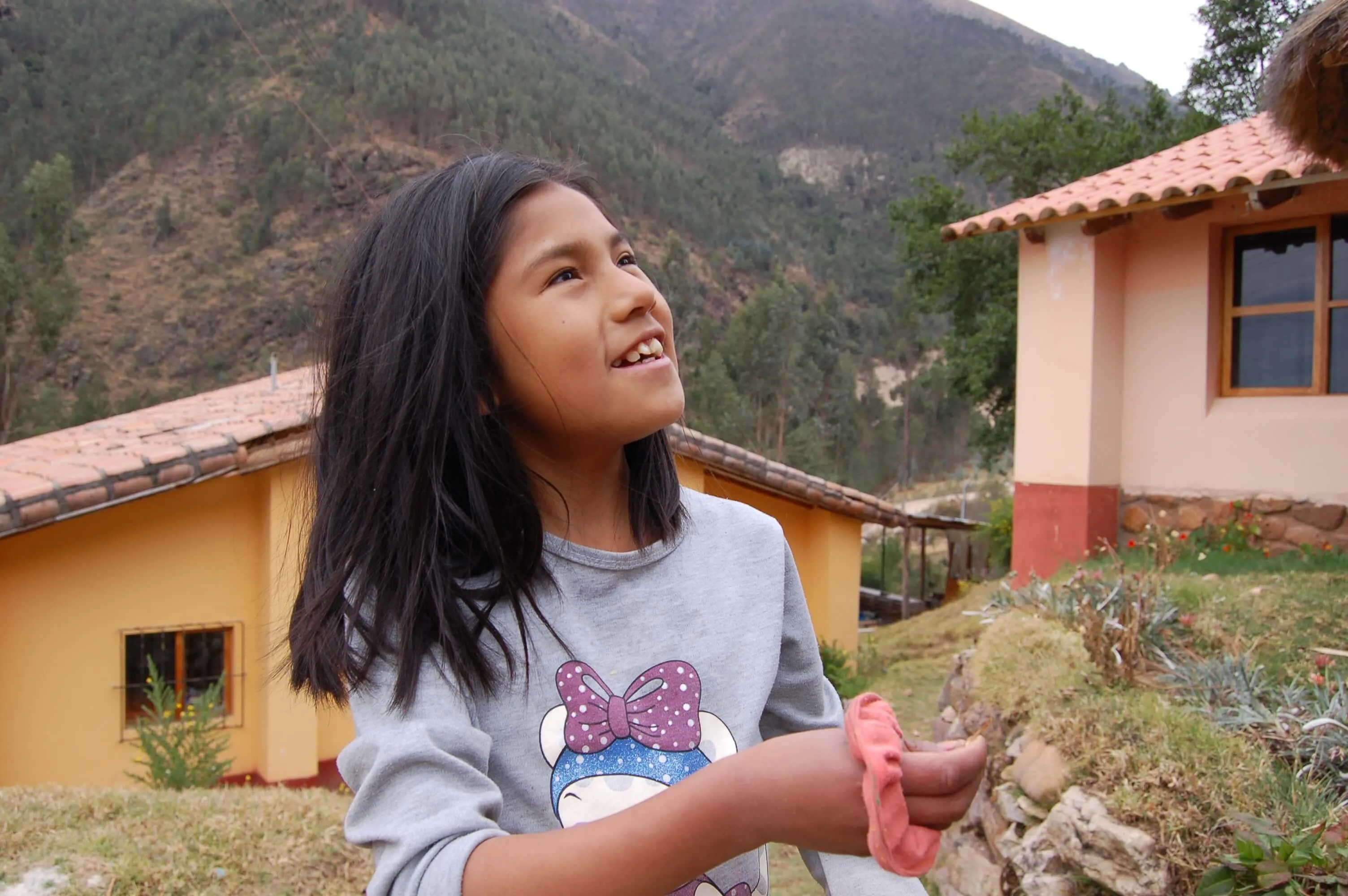 Young girl in a grey sweatshirt smiling joyfully against a backdrop of mountains and terracotta-colored houses in Peru.