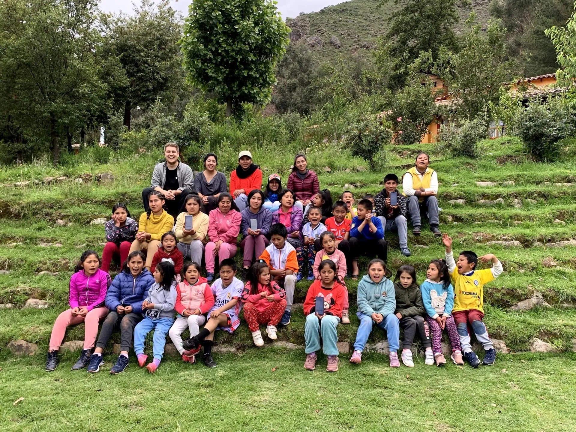 Group photo of Peruvian children and teachers sitting on a grassy hillside, wearing colorful casual clothes, with mountains in the background.