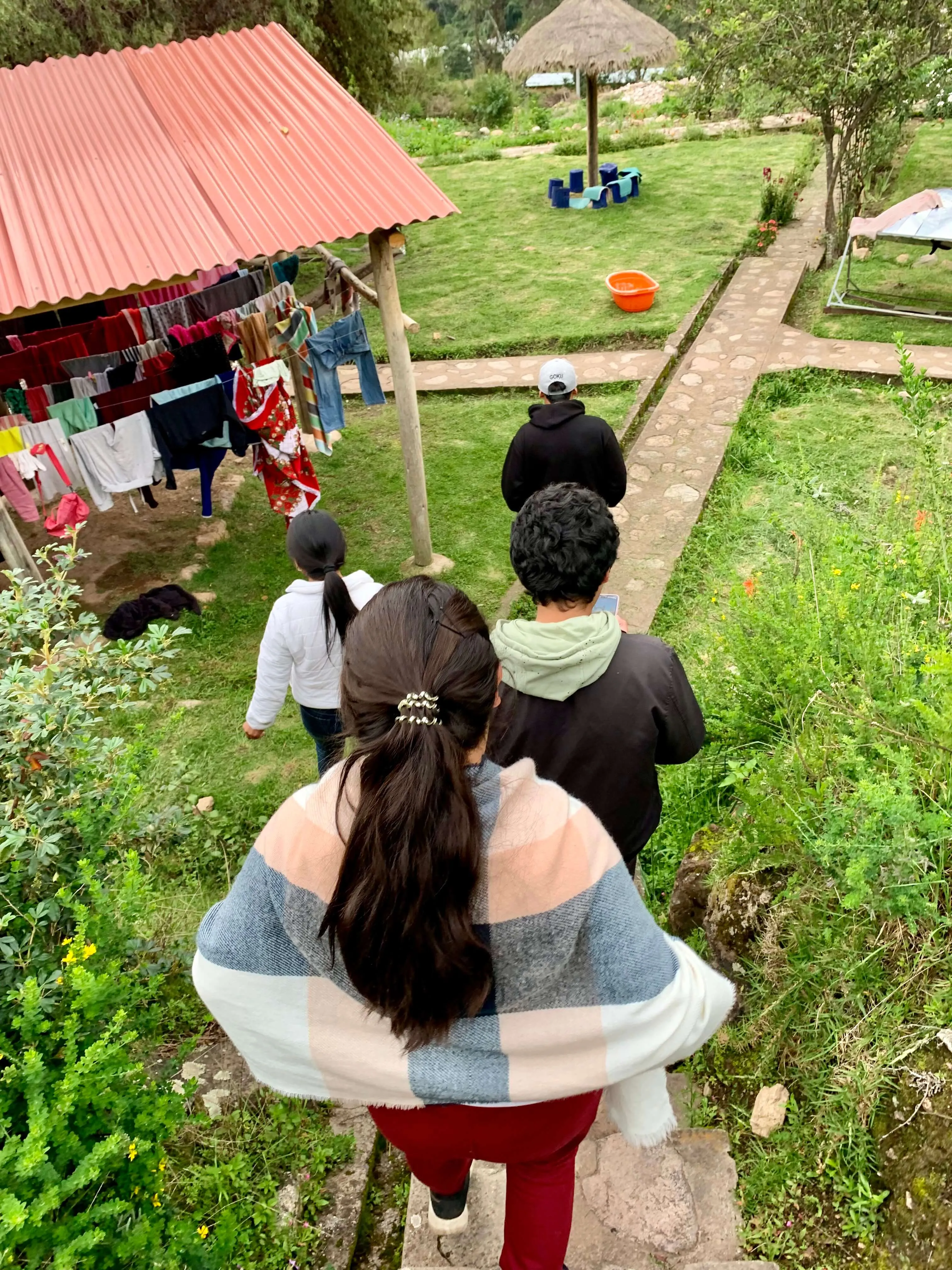 View down stone pathway of a garden area with clothesline under pink metal roof, people walking down path, and mountain landscape visible.